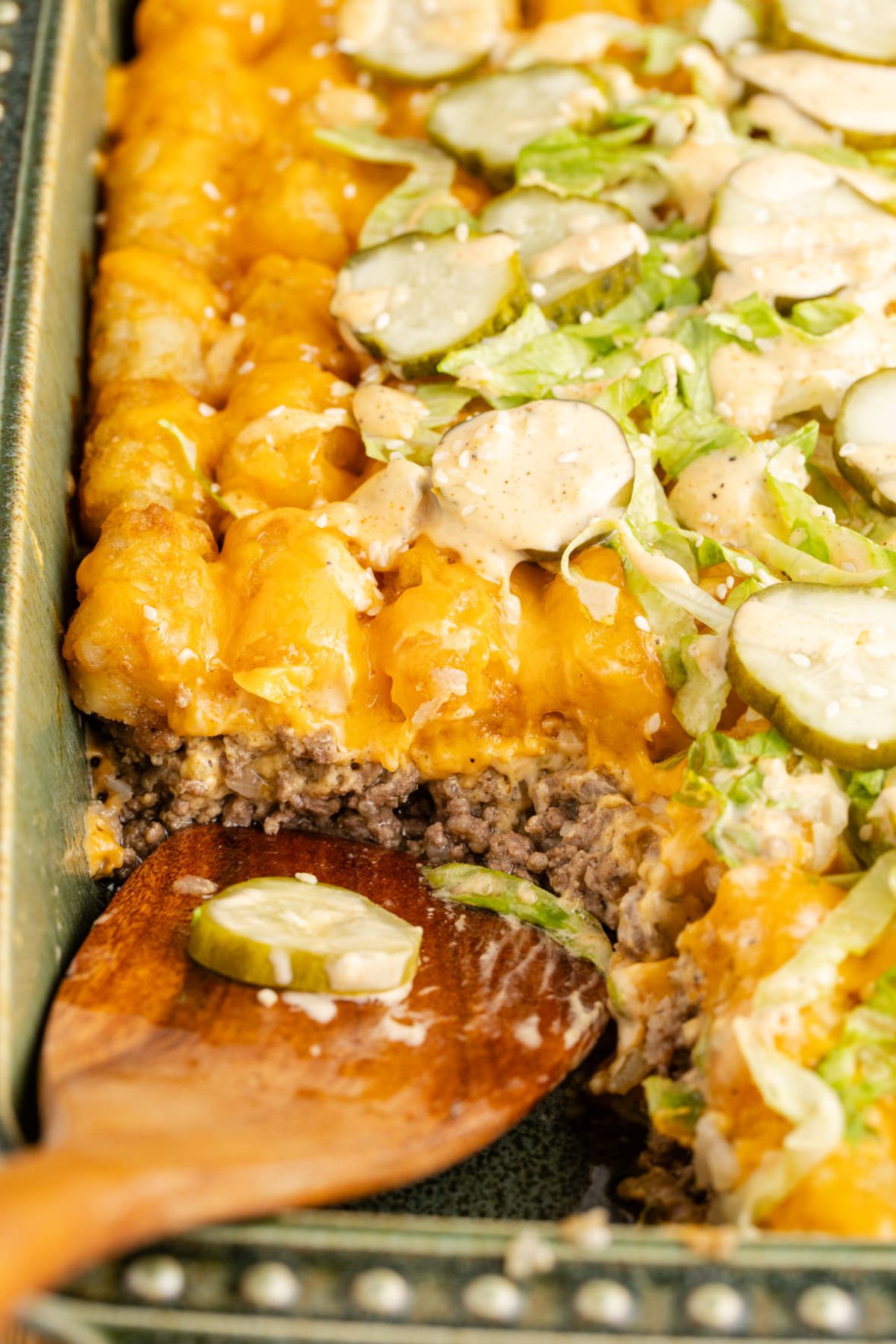 Beef, tater tots, cheese, lettuce, pickles and sauce on top being scooped with a wooden spoon.