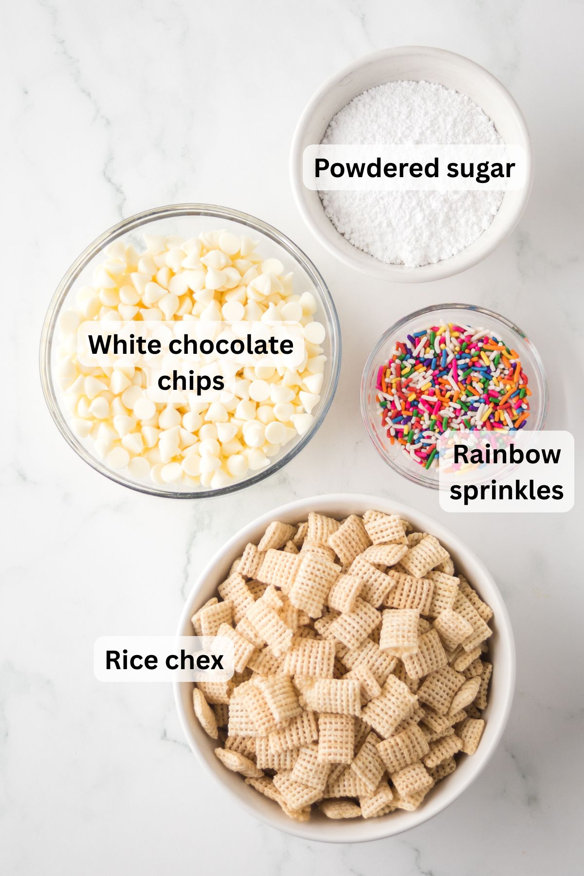 The ingredients needed to make birthday cake Chex mix in bowls on the table with labels to identify each one.