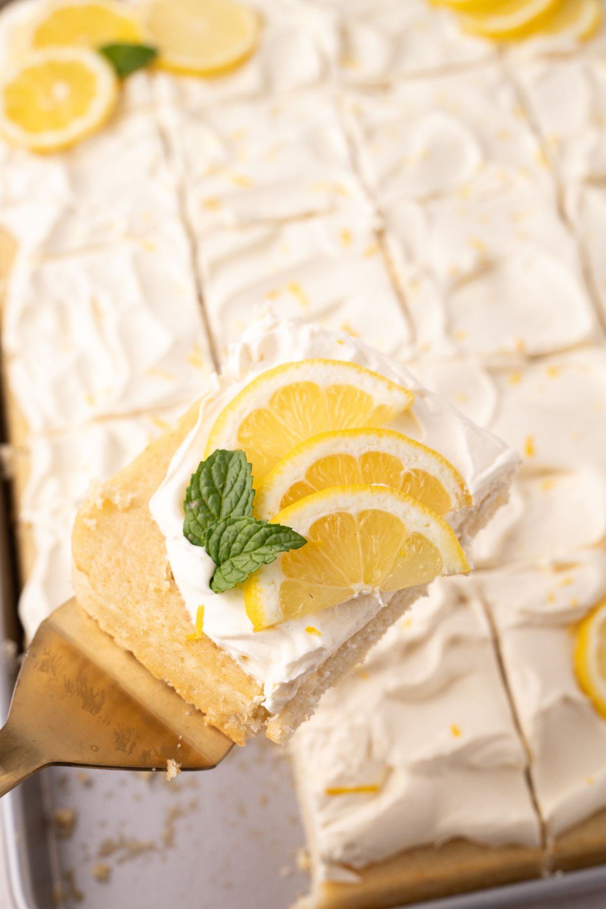 A slice of lemon cake being held up in the air above a sheet pan of cake.