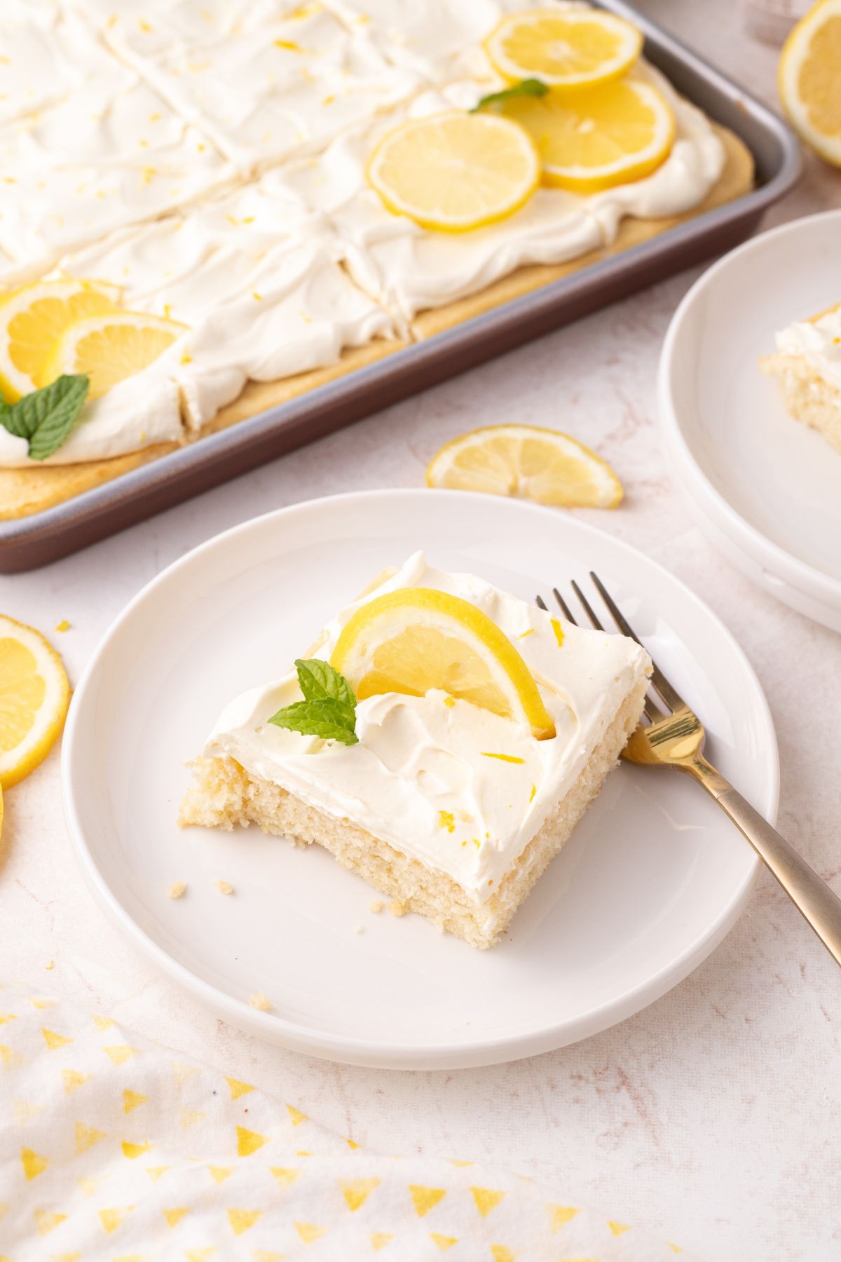 A slice of lemon sheet cake with a lemon slice and mint on a plate with a fork.