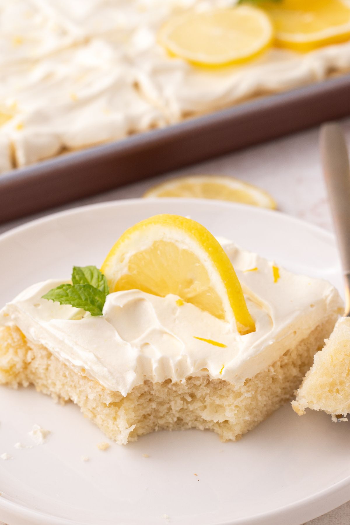 A lemon sheet cake slice with a piece missing and a lemon wedge on top.