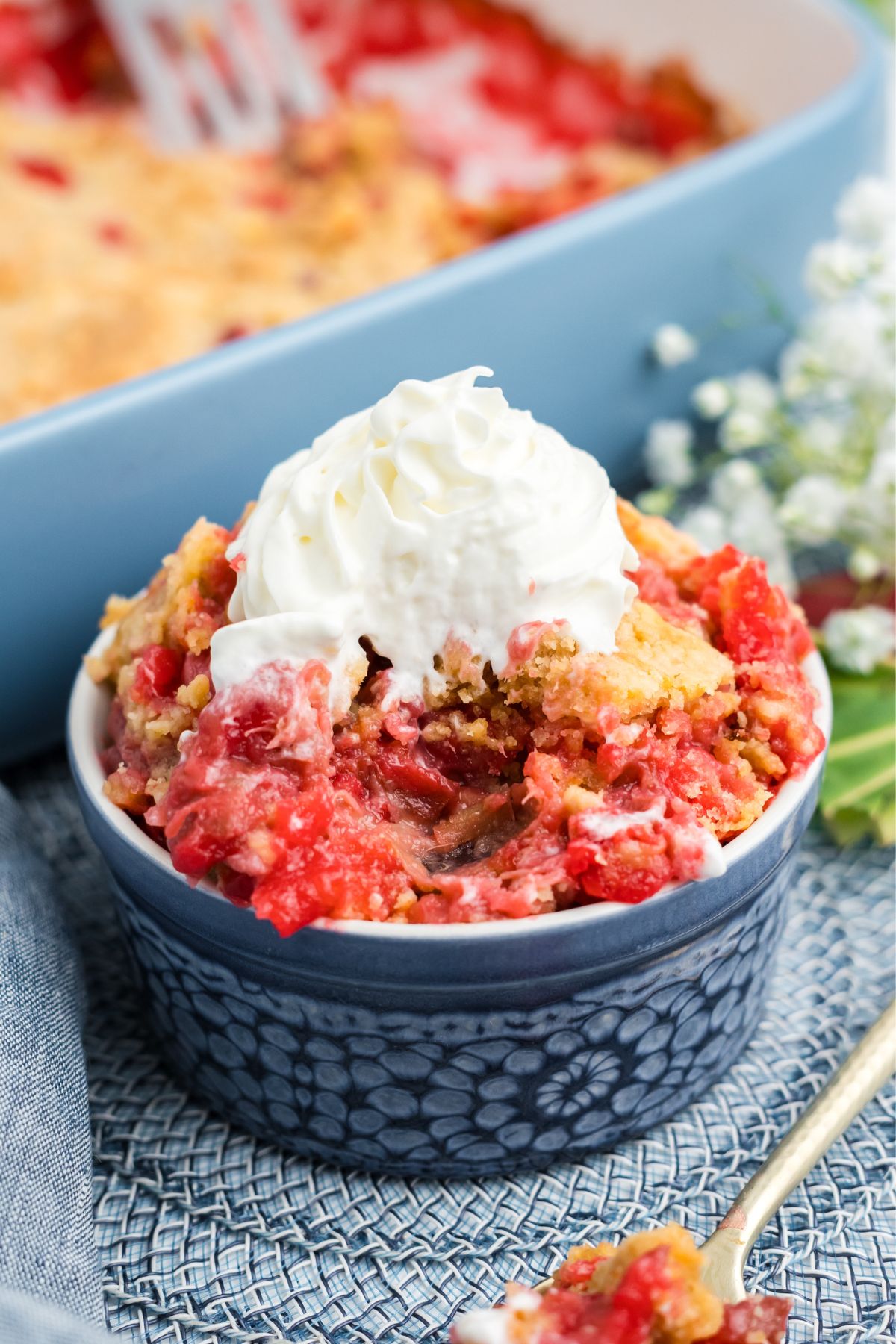 A ramekin with a serving of rhubarb cake topped with whipped cream and a spoon in the front with some cake on it.