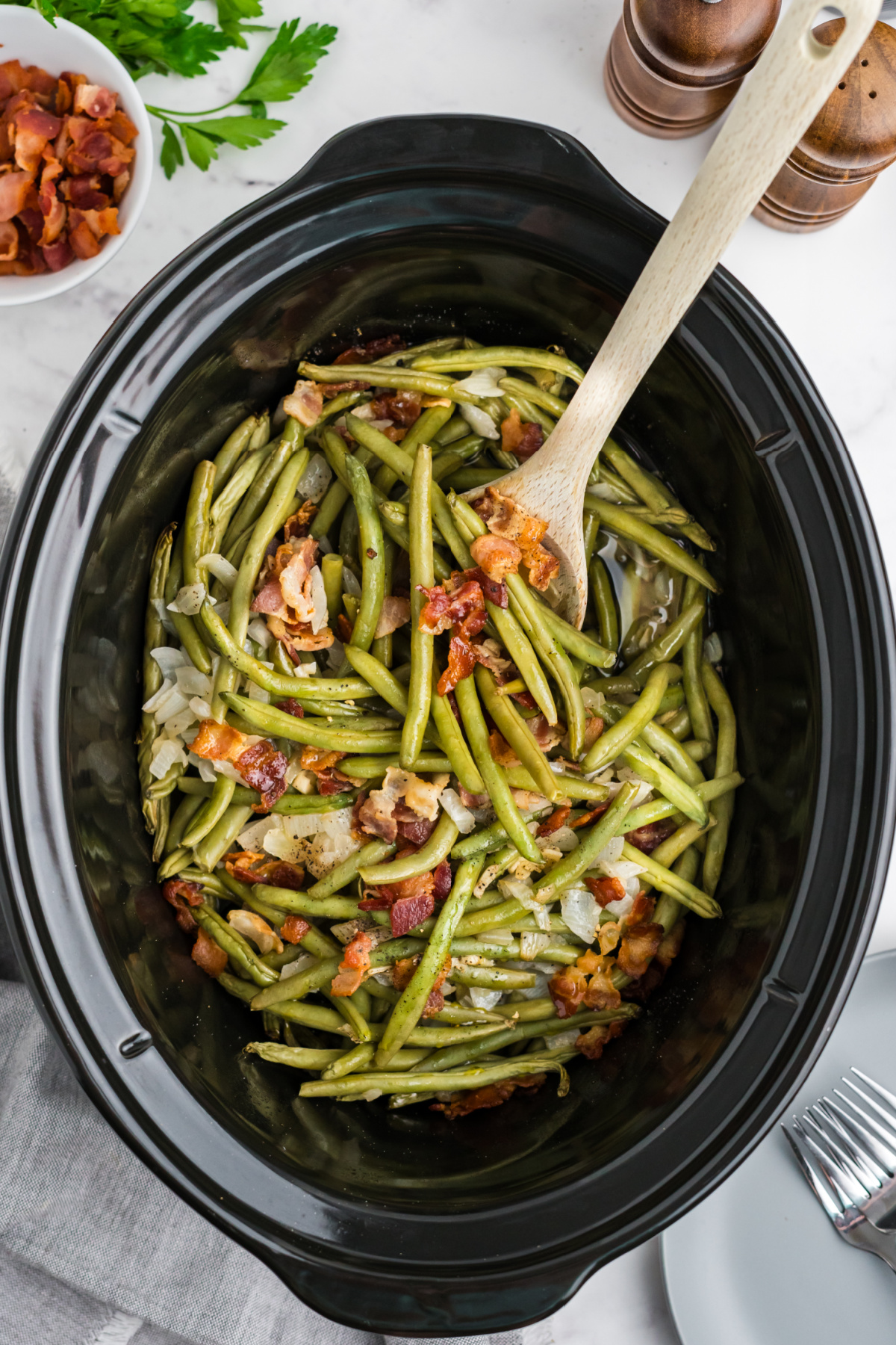 Green beans and bacon in the slow cooker with a wooden spoon mixing.