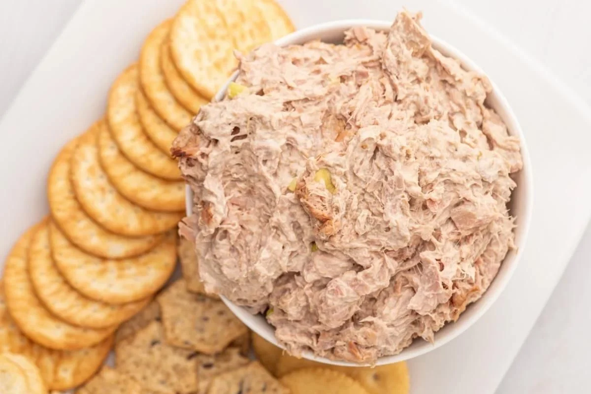 A bowl of tuna salad served with a variety of round crackers and snack dips on a white plate.