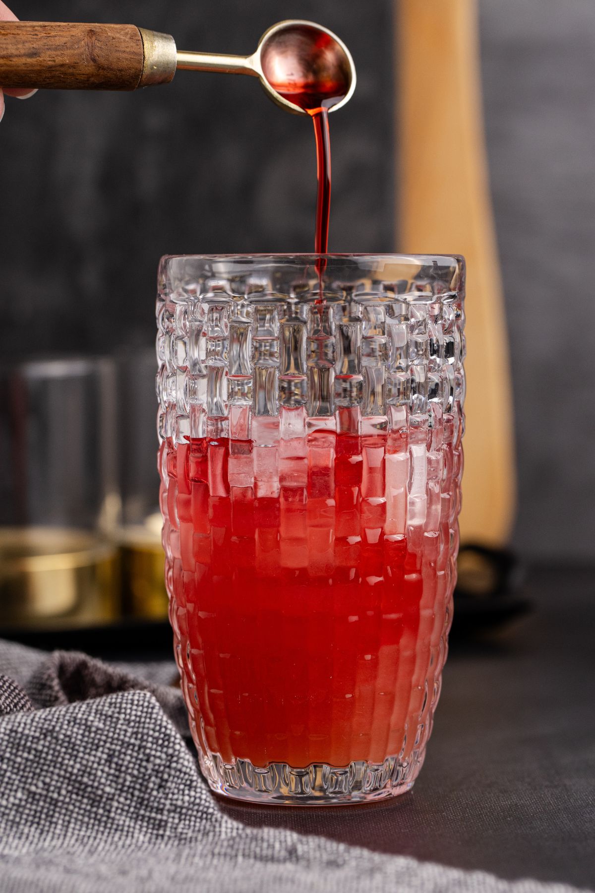 Grenadine being poured into shaker halfway filled with ice, vodka, cranberry juice and pineapple juice.