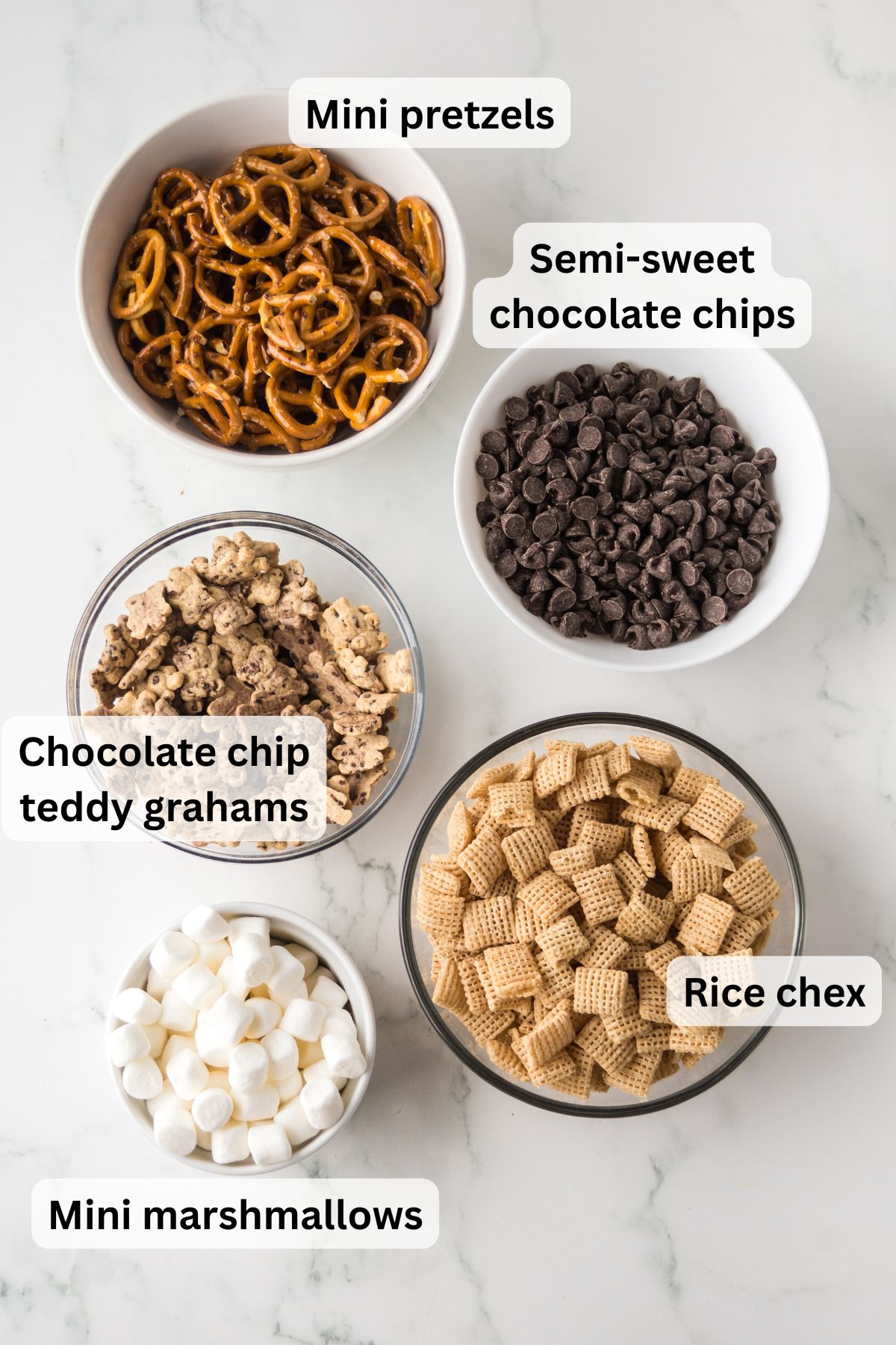 The ingredients needed to make s'mores snack mix in bowls on the table with labels to identify each one.