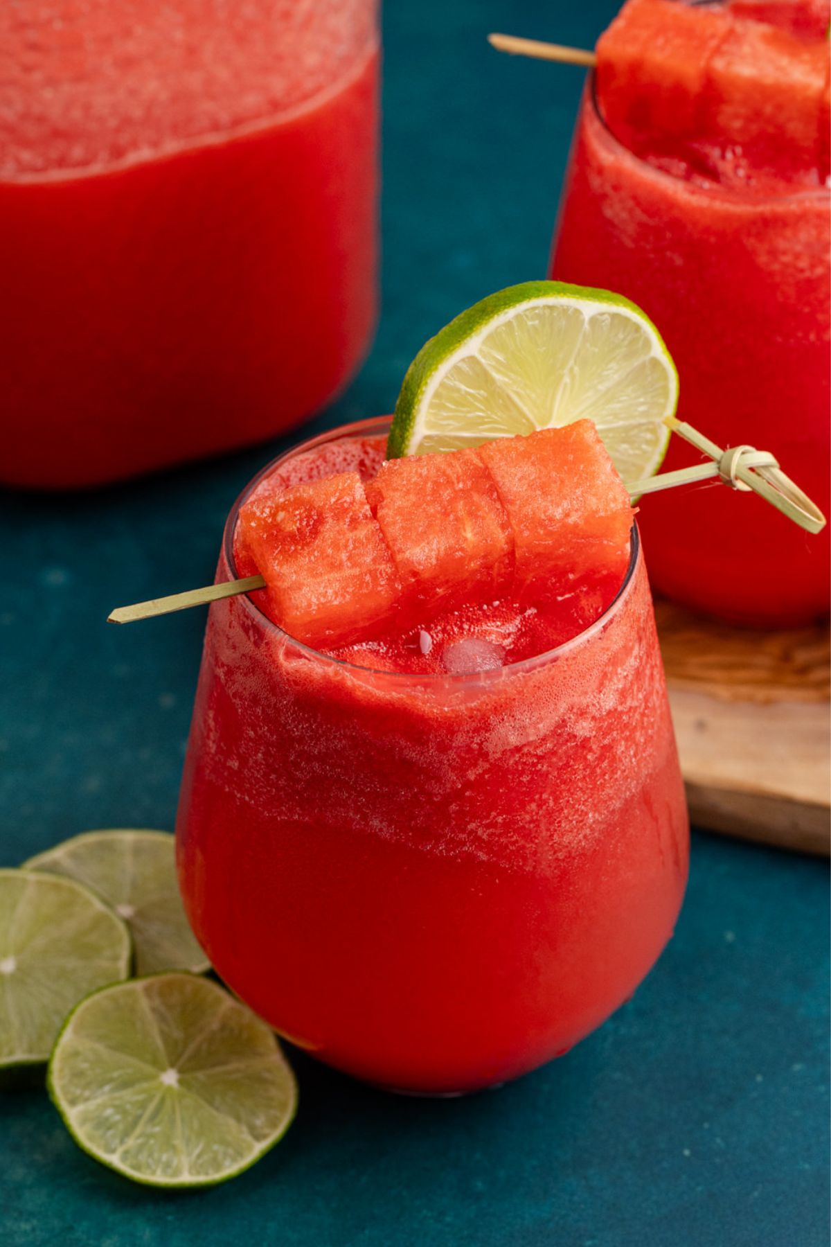Cocktail garnished with a lime slice and watermelon cubes beside pitcher.