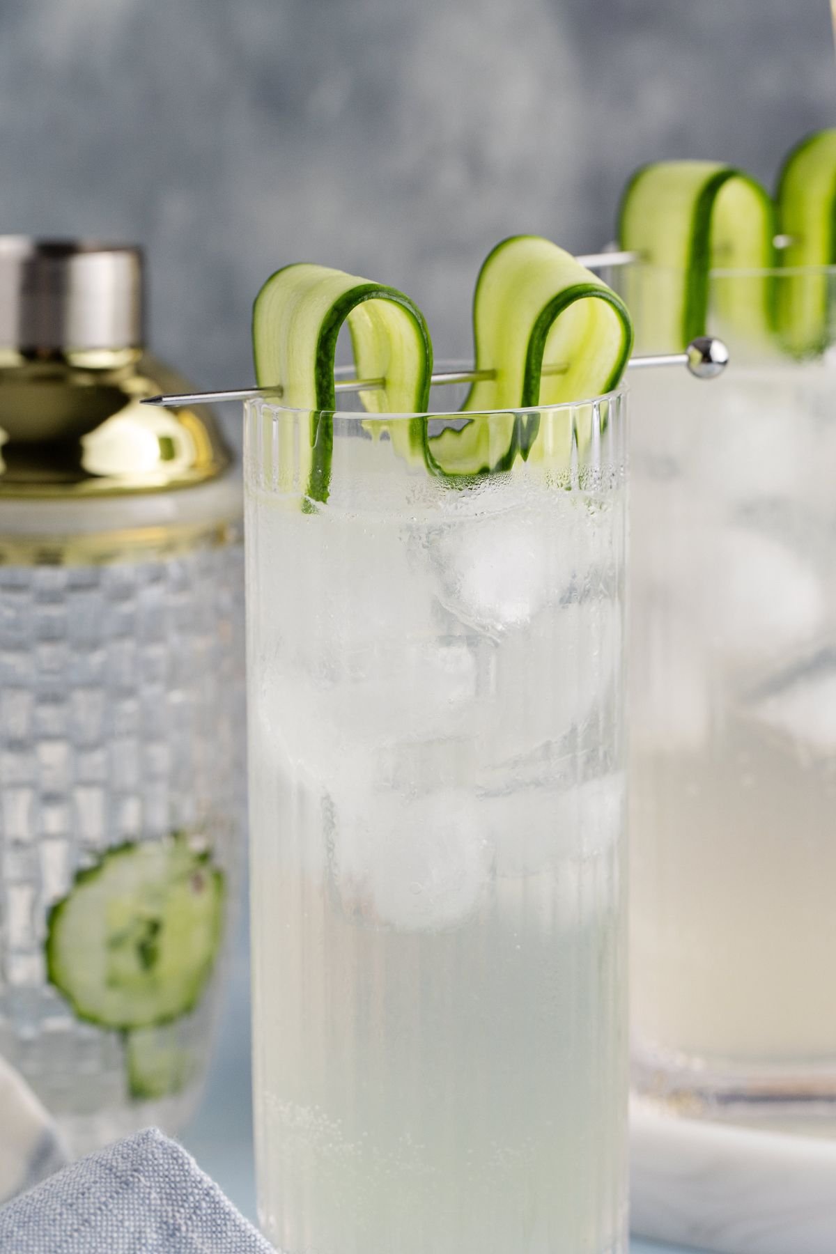 Two cocktails garnished with cucumber a slice in a table with the shaker.