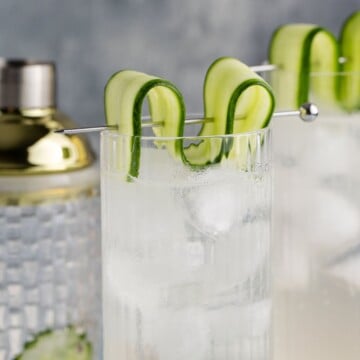 White linen cocktail garnished with cucumber on table with cocktail shaker and another drink in background.