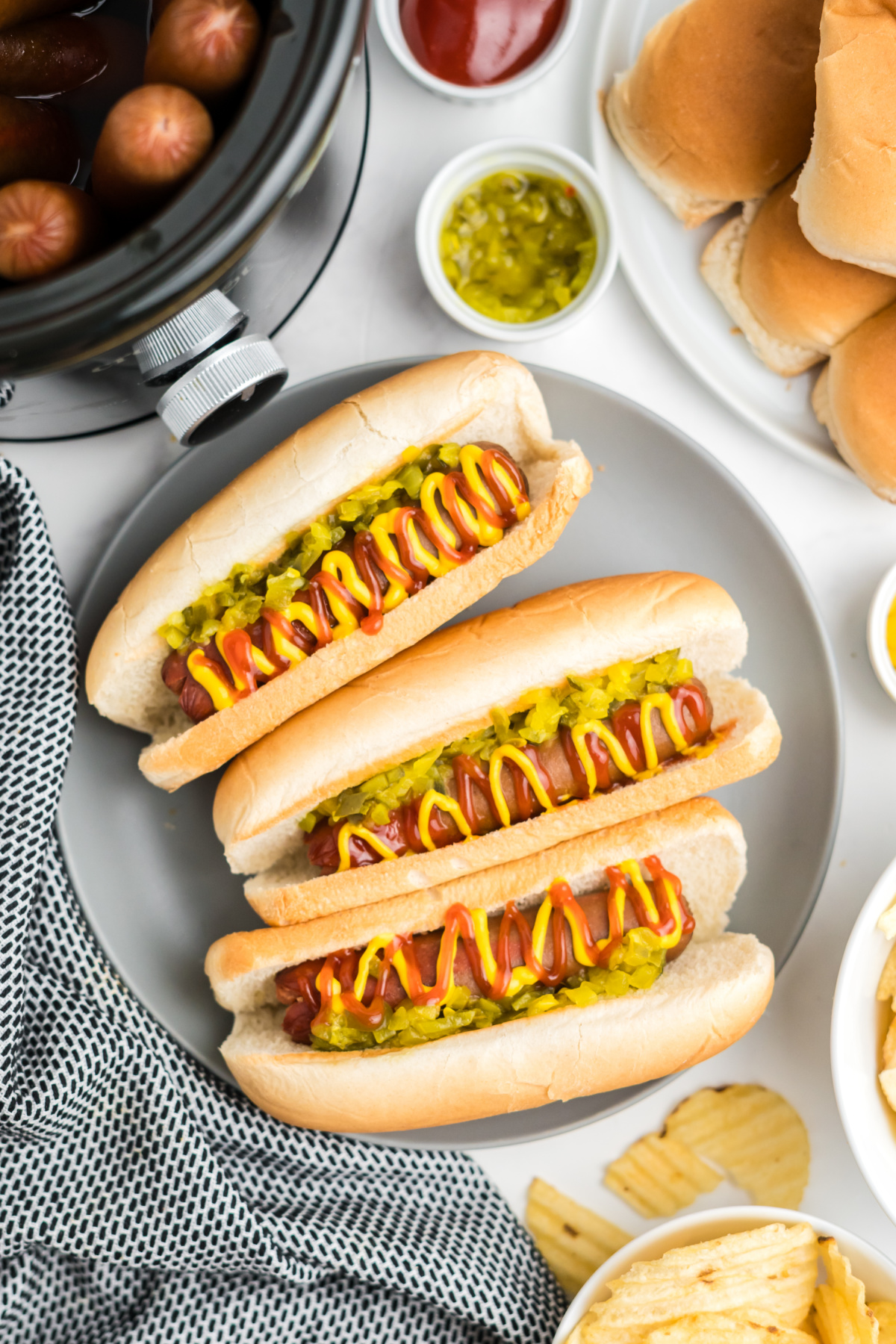 Three hot dogs on a plate with relish, mustard and ketchup.