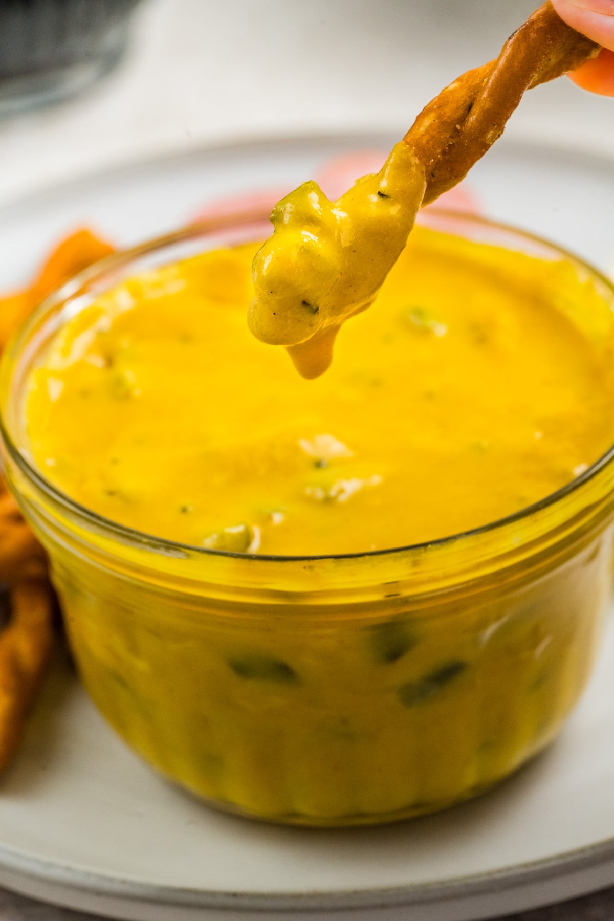 A jar of mustard with a pretzel lifted out of it dripping after being dipped.