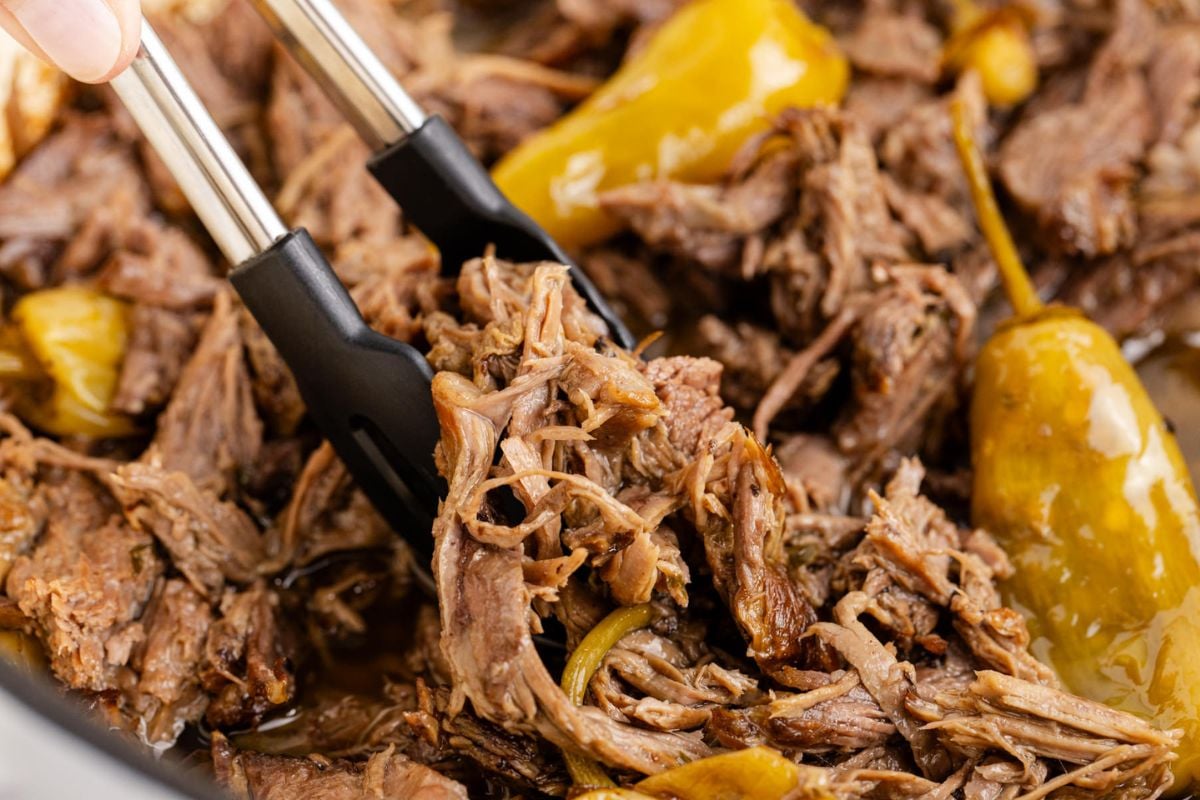 A pile of shredded pot roast with tongs squeezing a portion.