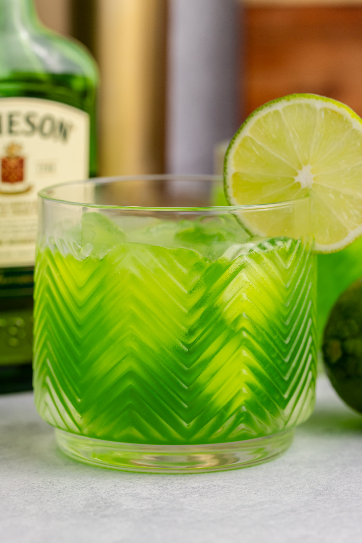 A glass with whiskey, syrup, citrus, and green food coloring with a lime garnish.