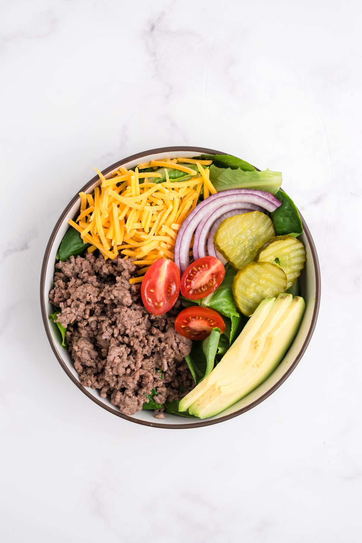 Completed burger bowl including shredded cheese, lettuce, sliced onion, pickles, tomato and avocado.