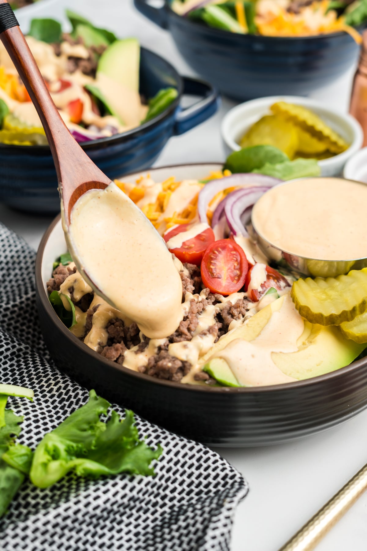 A wooden spoon drizzling creamy burger bowl sauce on to of ground beef in a burger bowl.