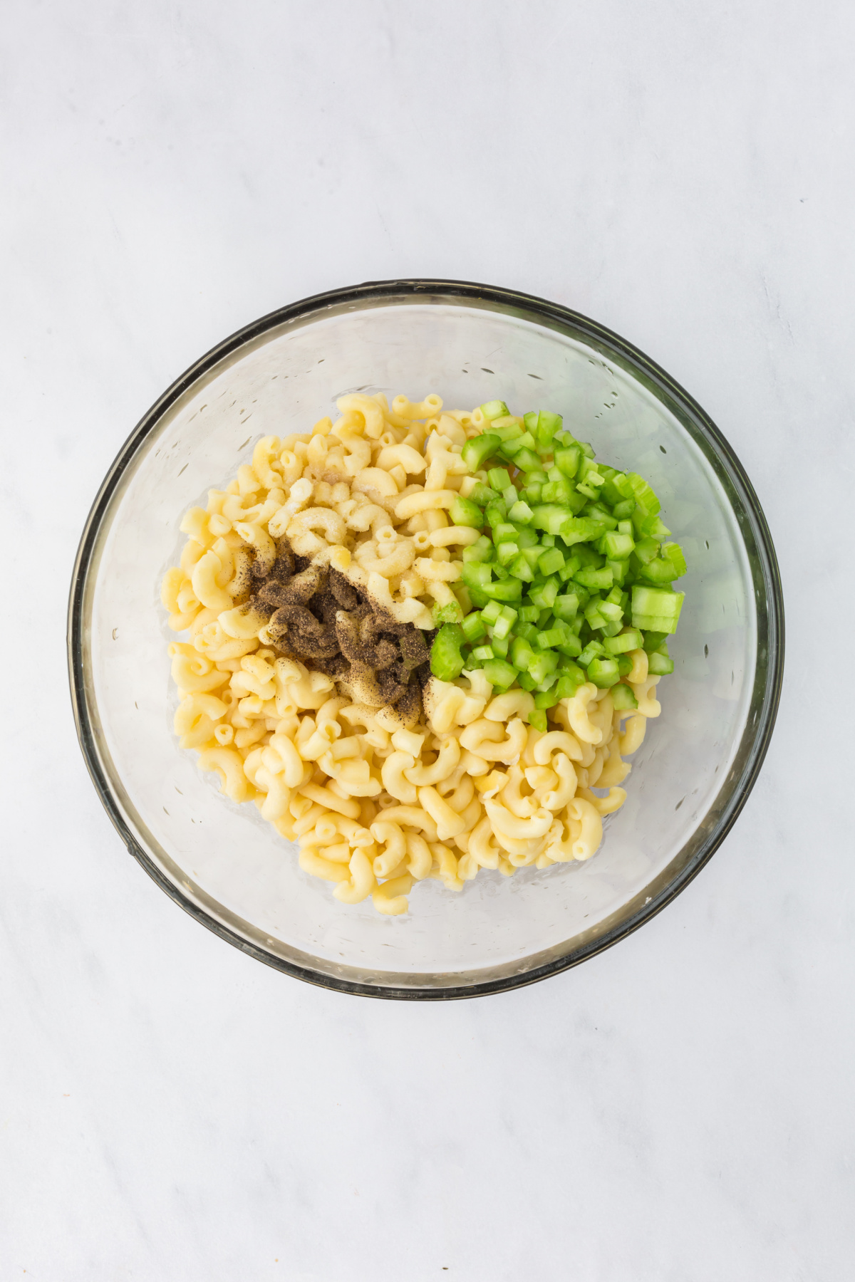 Celery, salt and pepper are being added to bowlful of macaroni on a counter.