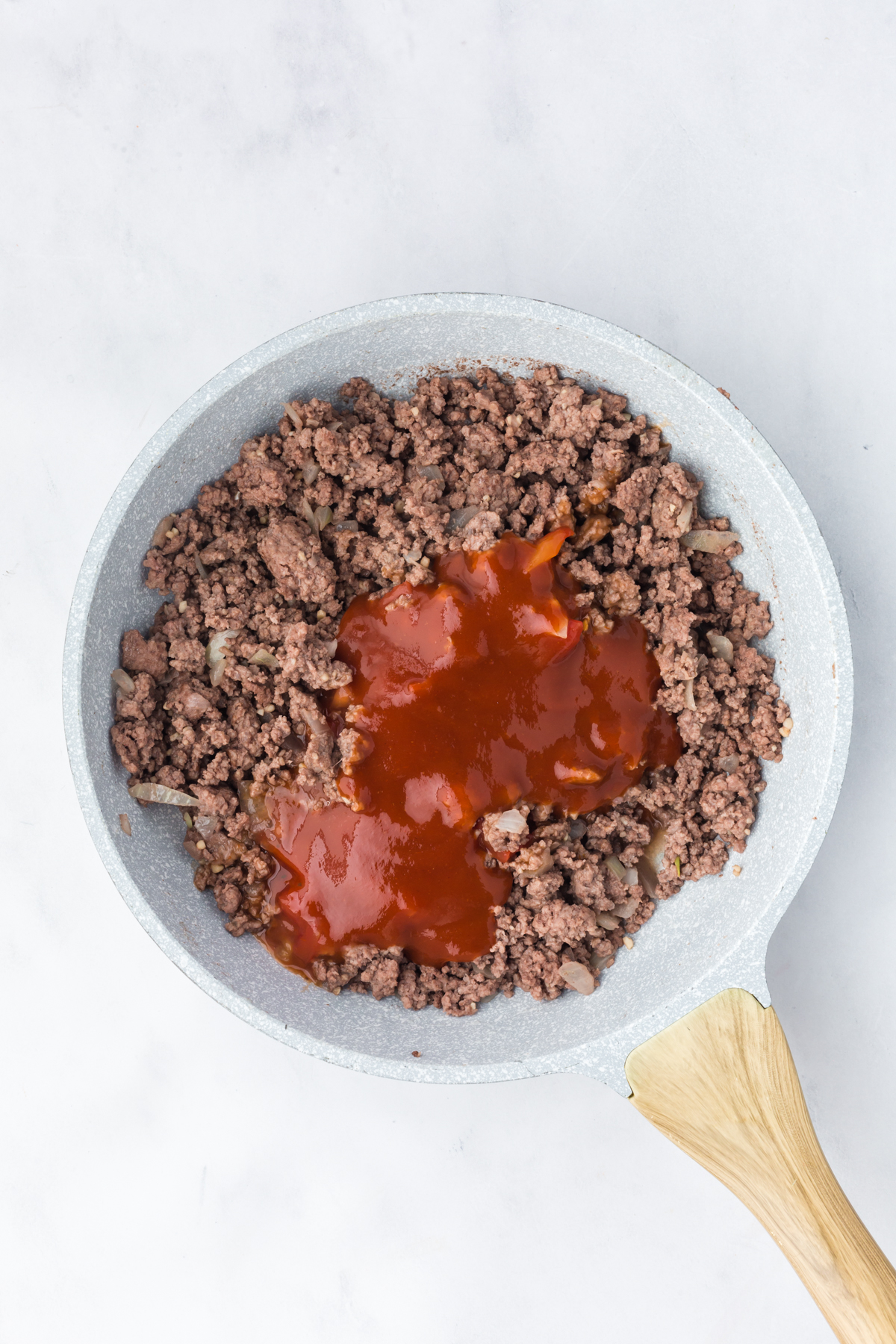 Sloppy joe sauce being added to cooked ground beef in skillet.