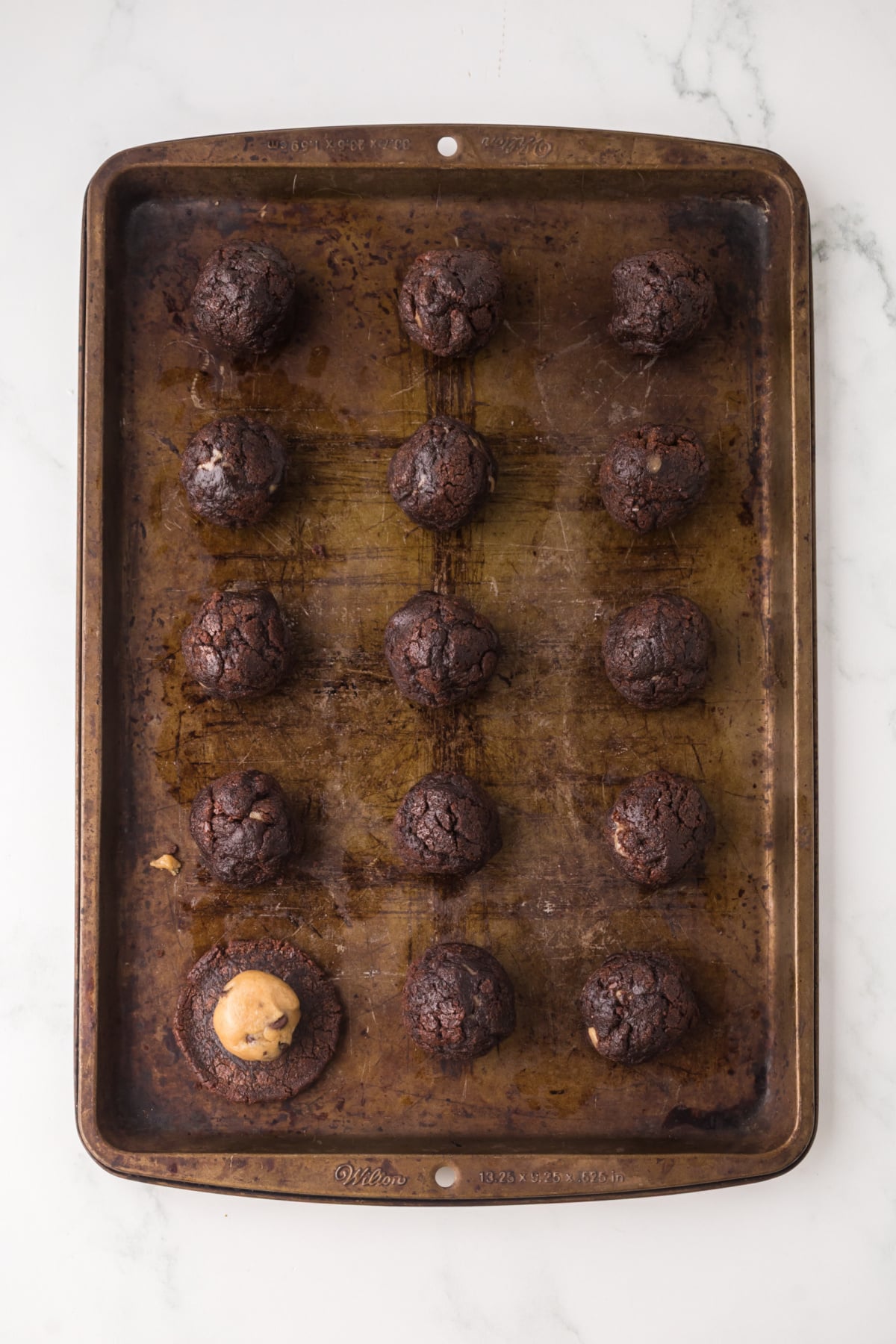 A baking sheet with one cookie dough ball in top of of flattened brownie bite surrounded by rolled cookie dough balls.