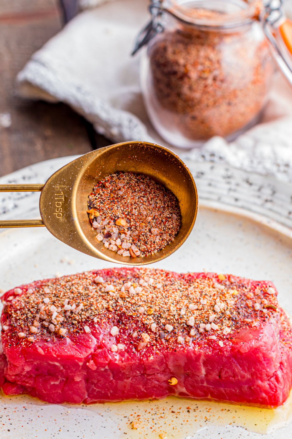 A raw steak on a plate being coated in steak seasoning.