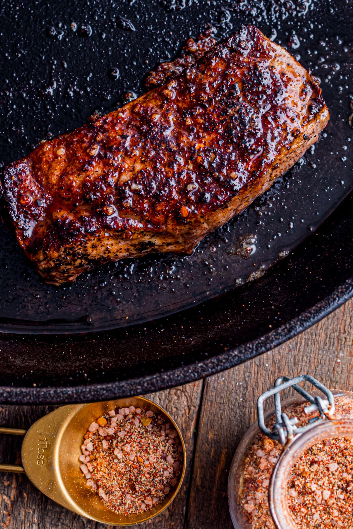 A cooked steak in a pan with a wonderful crust and steak seasoning in jar and spoon beside it.
