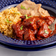 Asado de puerco on a blue plate with a side of rice and refried beans.