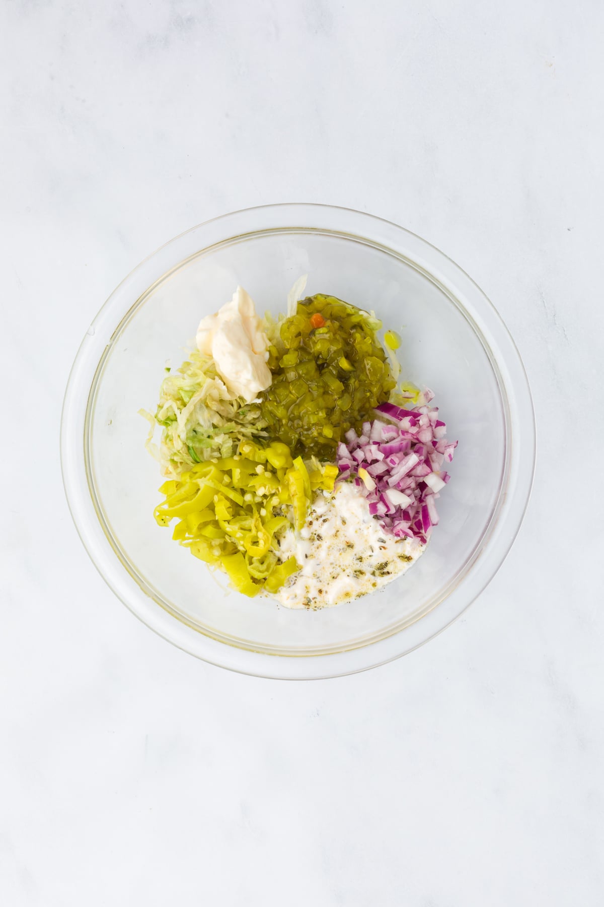 A bowl with shredded lettuce, mayonnaise, red onion, relish, banana peppers, and submarine dressing.