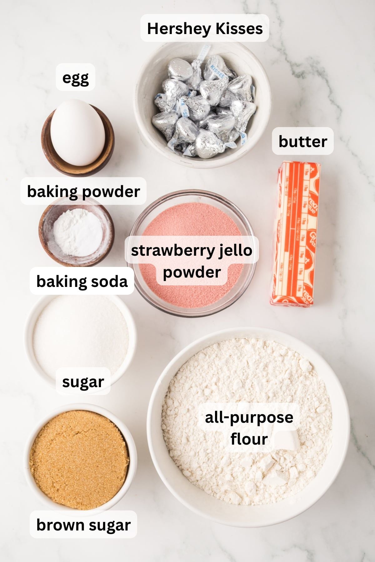 All the ingredients needed to make Cookies With Hershey Kisses (Strawberry Flavor) including butter sugar flour strawberry jello and chocolate kisses in bowls on a table. 
