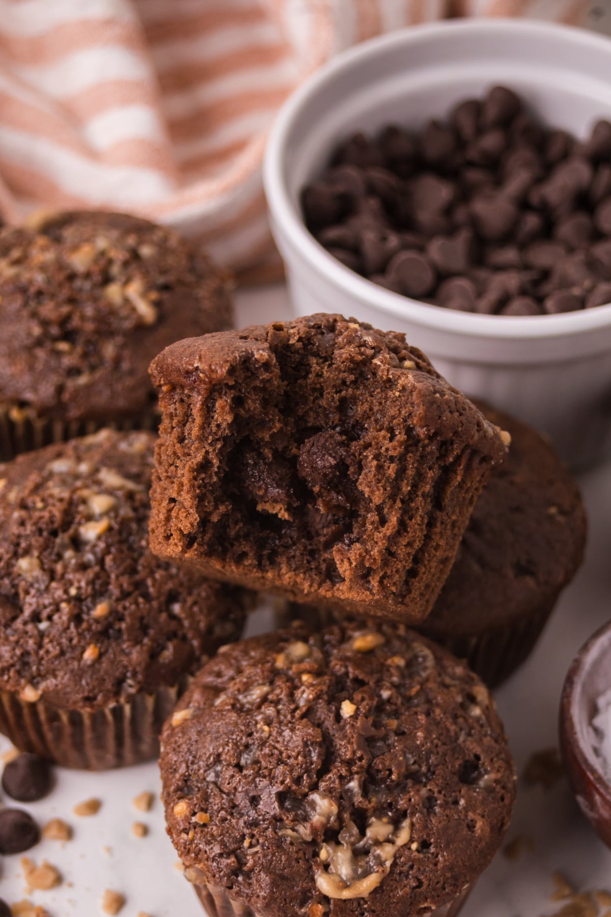 Freshly baked double chocolate chip muffins with domed tops and melted chocolate chips scattered on a table with one on top missing a bite.