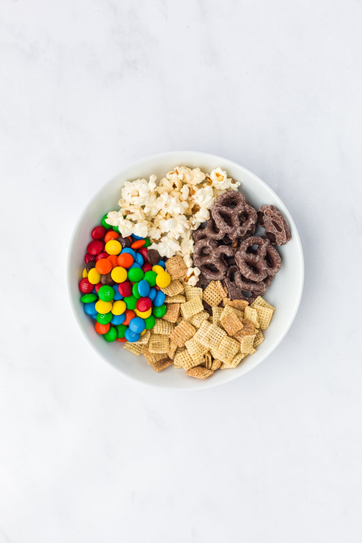 adding popcorn cereal chocolate and pretzels to a mixing bowl for trail mix