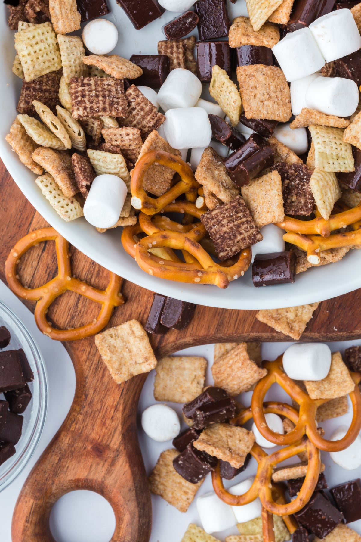 trail mix in a bowl on a cutting board surrounded by cereal pretzels marshmallows and chocolate on the table