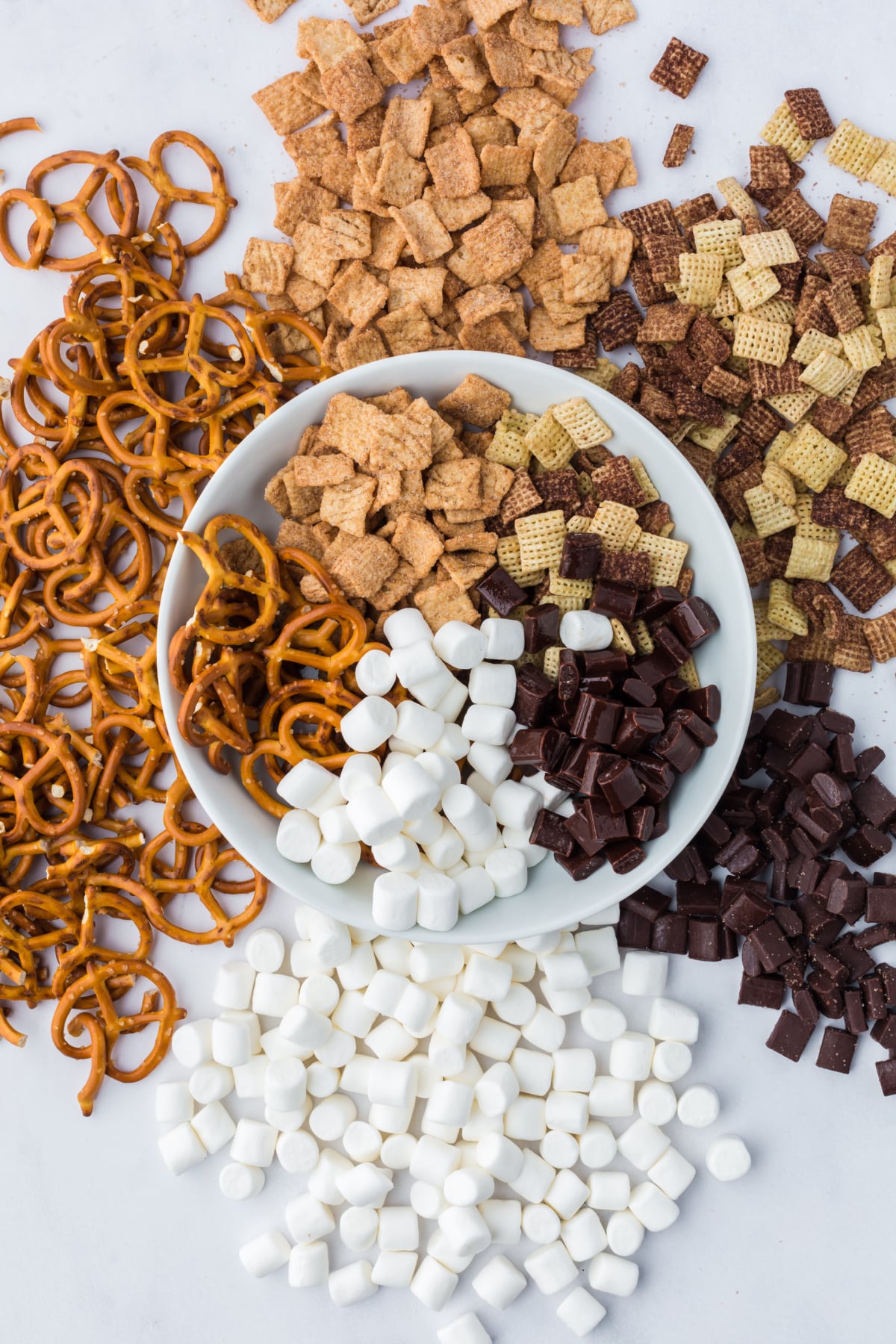 Ingredients needed to make s'mores trail mix in a bowl surrounded by ingredients on a table