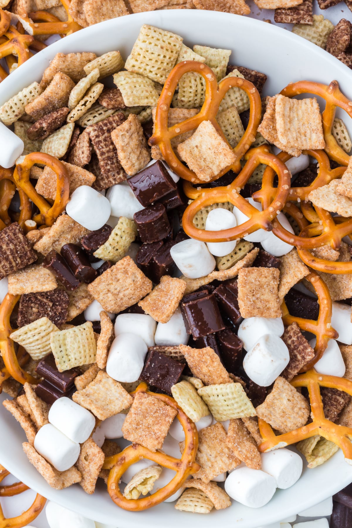 trail mix in a bowl surrounded by cereal pretzels marshmallows and chocolate on the table