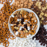 s’mores trail mix in a bowl surrounded by cereal pretzels marshmallows and chocolate on the table