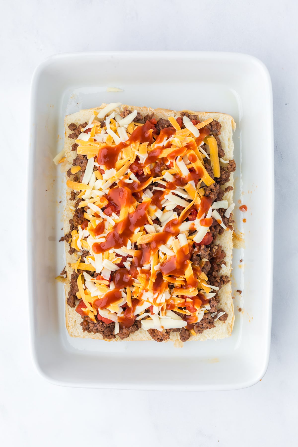 Layered ingredients over the bottom buns, starting with the ground beef, tomatoes, cheese, and taco sauce in casserole dish.