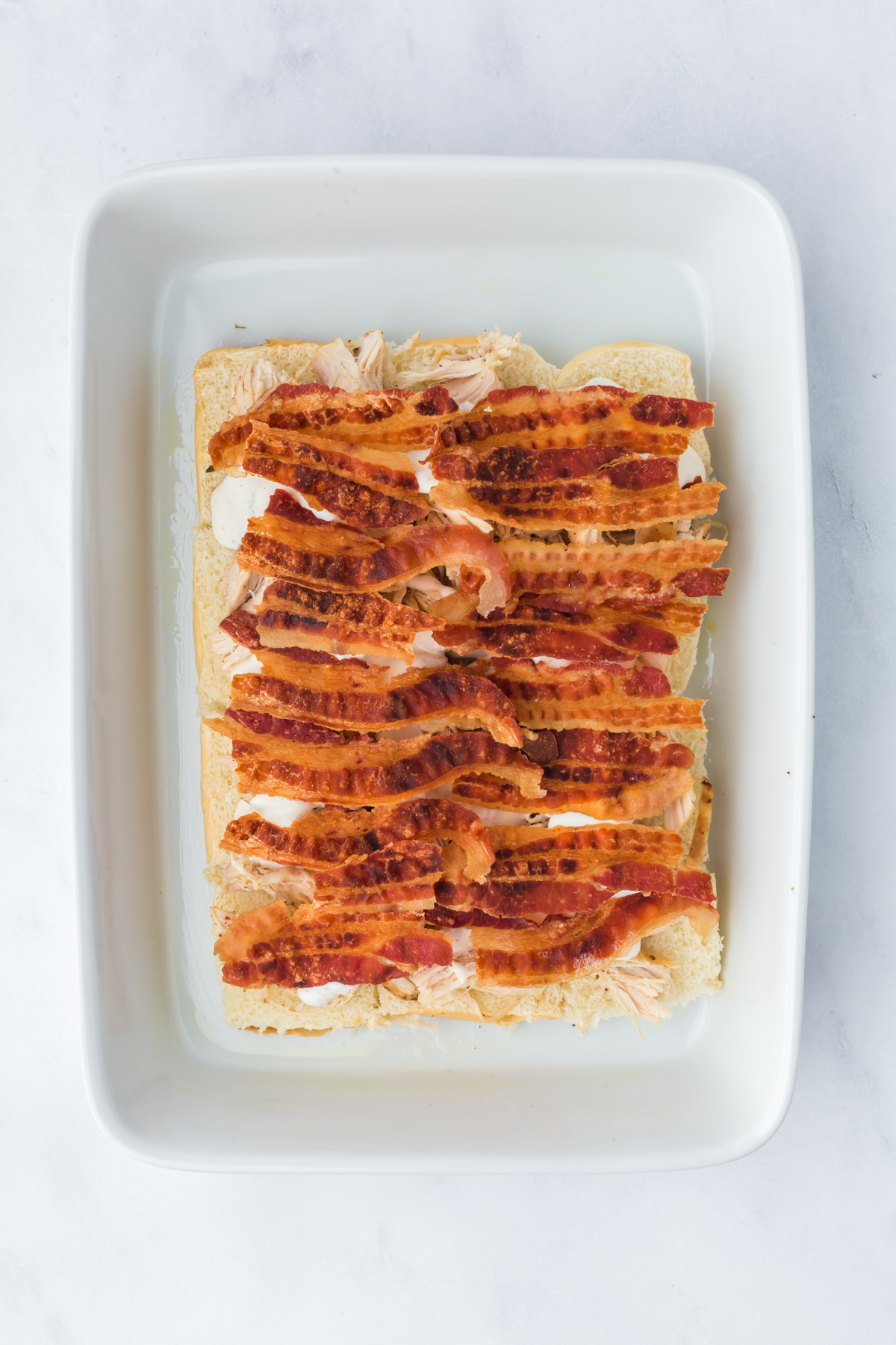 Toasted sliders in baking dish covered in cheese, chicken, drizzled ranch dressing and bacon slices.