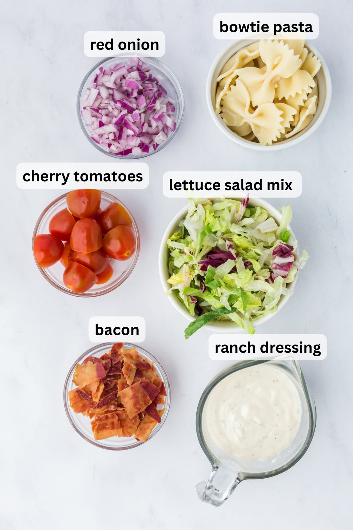 ingredients for bacon ranch dressing pasta salad including bacon lettuce tomatoes pasta and ranch dressing in bowls on a table