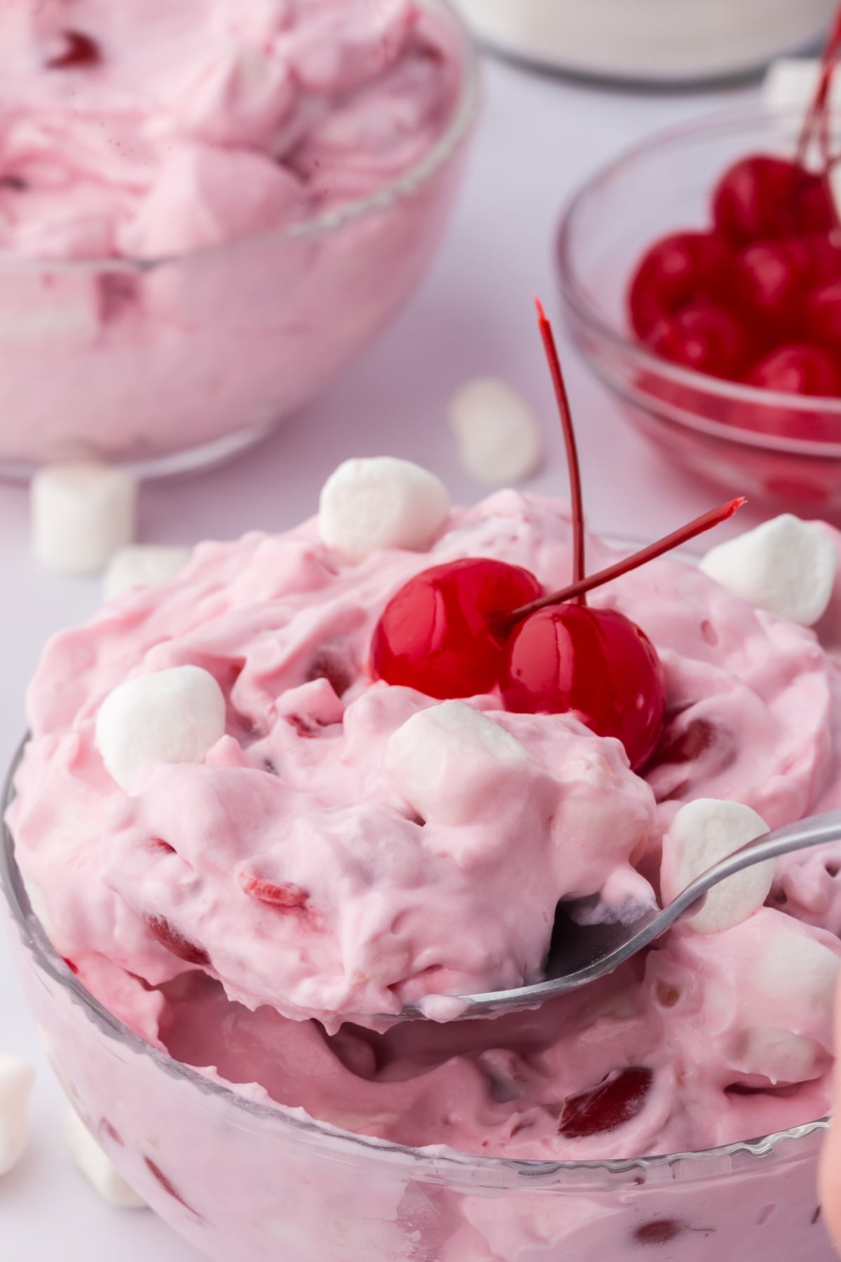 A bowl of cherry fluff topped with 2 cherries on a table with a spoon.