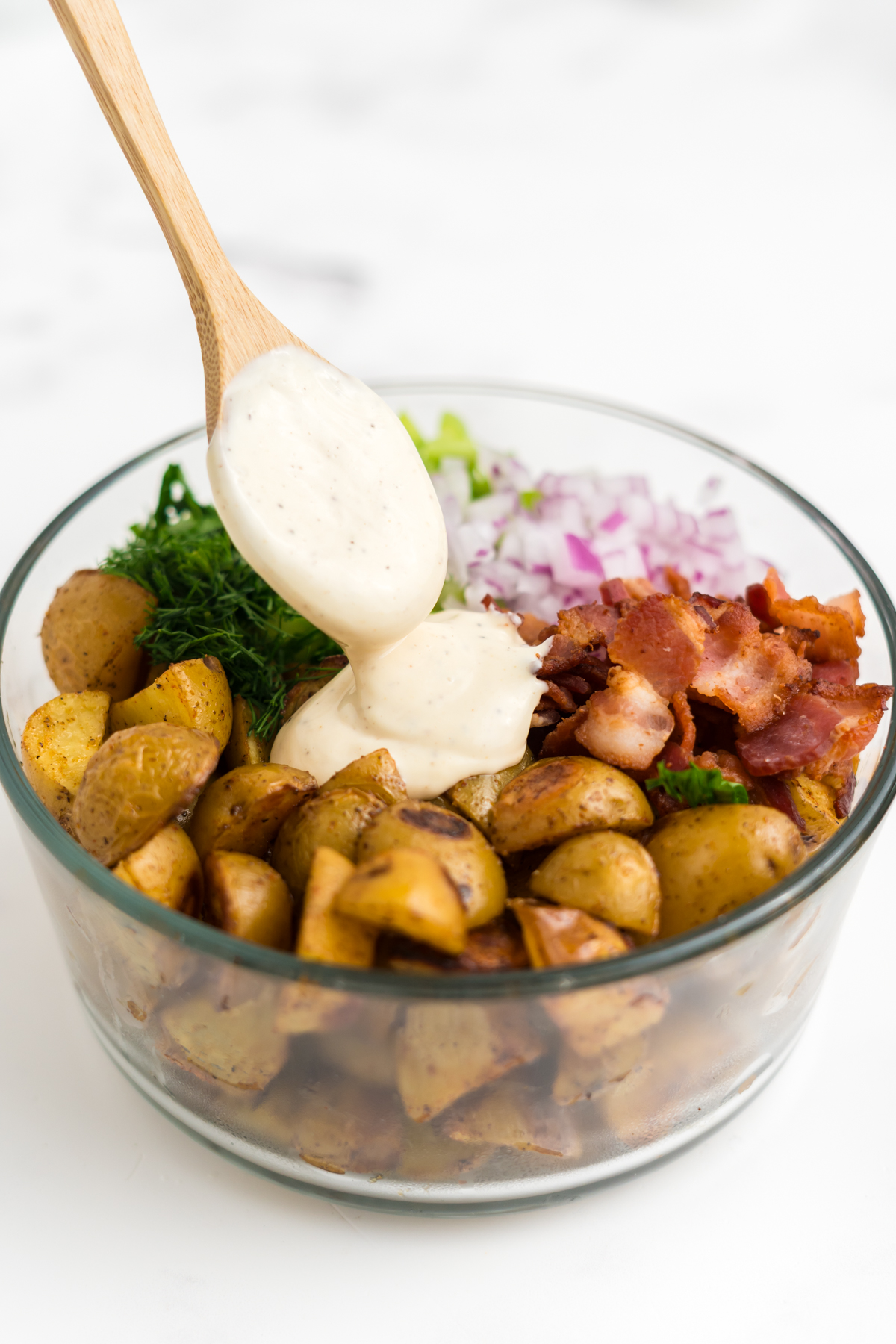 A bowl full of slightly cooled potatoes, celery, red onion, dill, parsley, and bacon with a wooden spoon adding dressing.