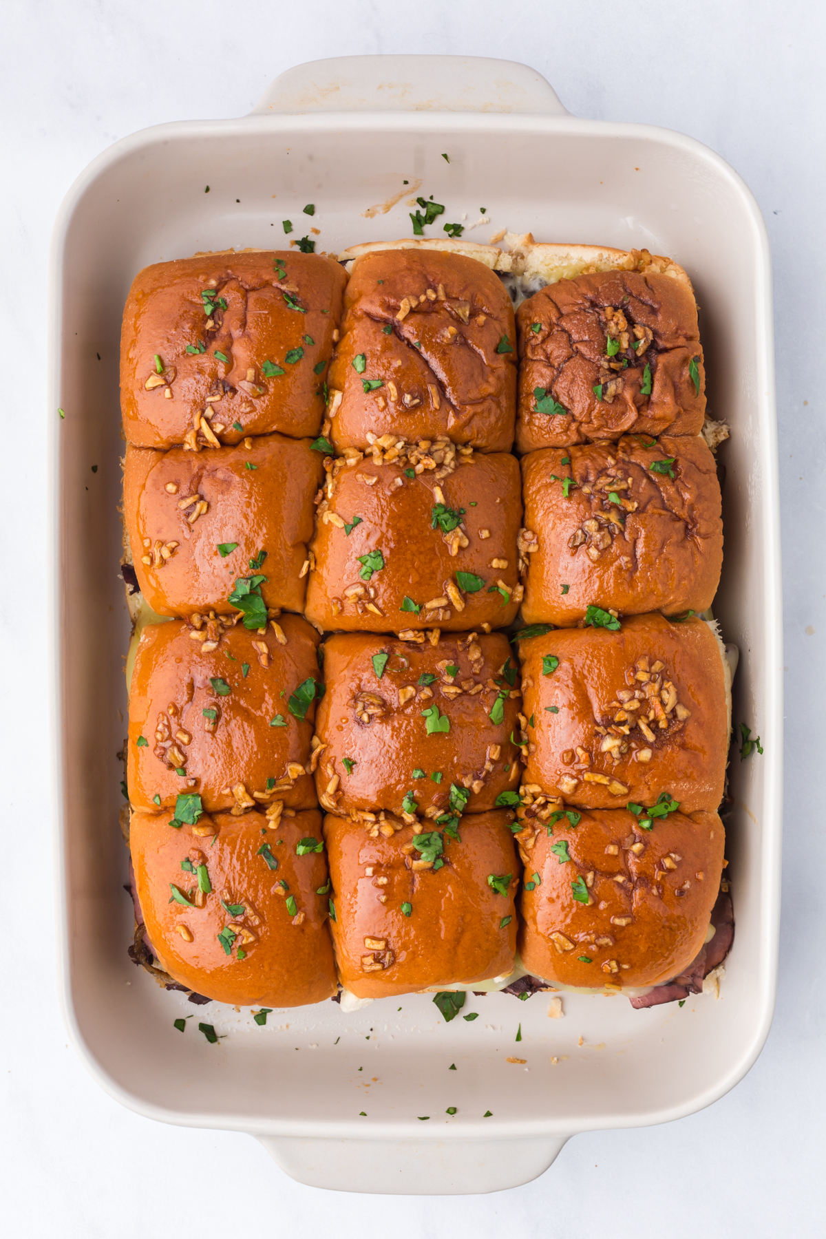 Baked sliders garnished with chopped parsley in casserole dish.