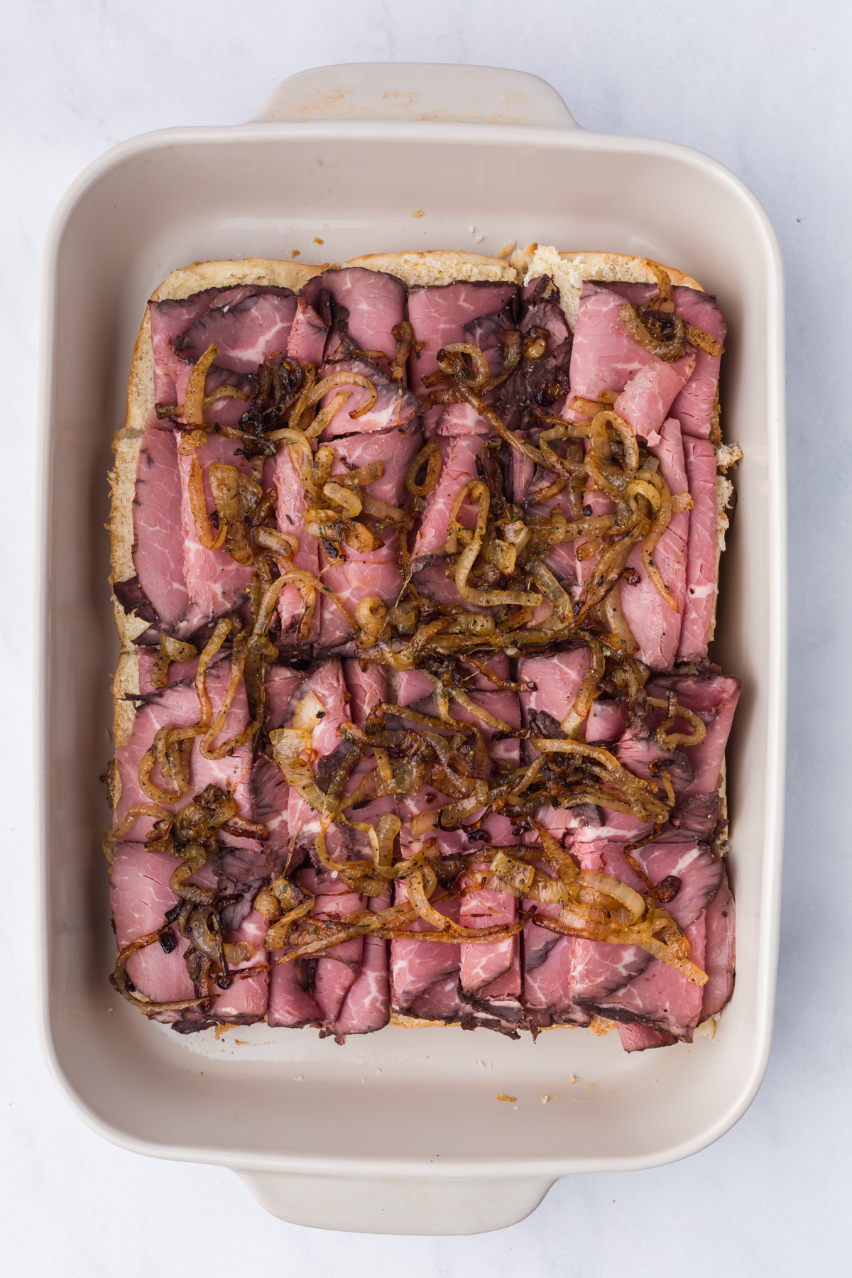 toasted slider bun bottom with cheese, roast beef, fried onions in casserole dish.