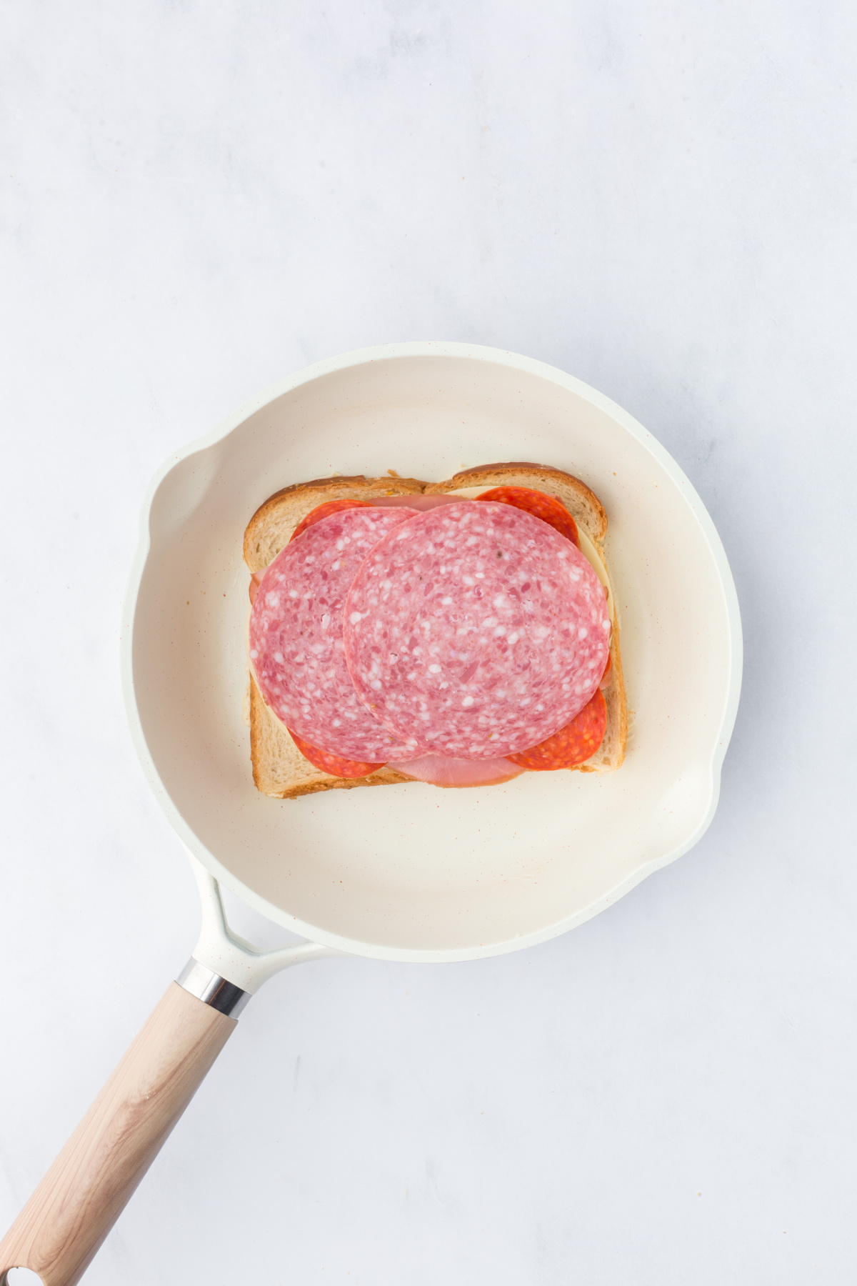 A slice of bread, butter side down, in skillet topped with provolone cheese, ham, pepperoni, and salami.