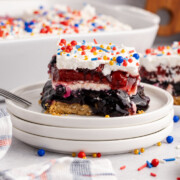 A slice of Red White and Blue Berry Dessert on a plate with a fork with dessert behind it on a table