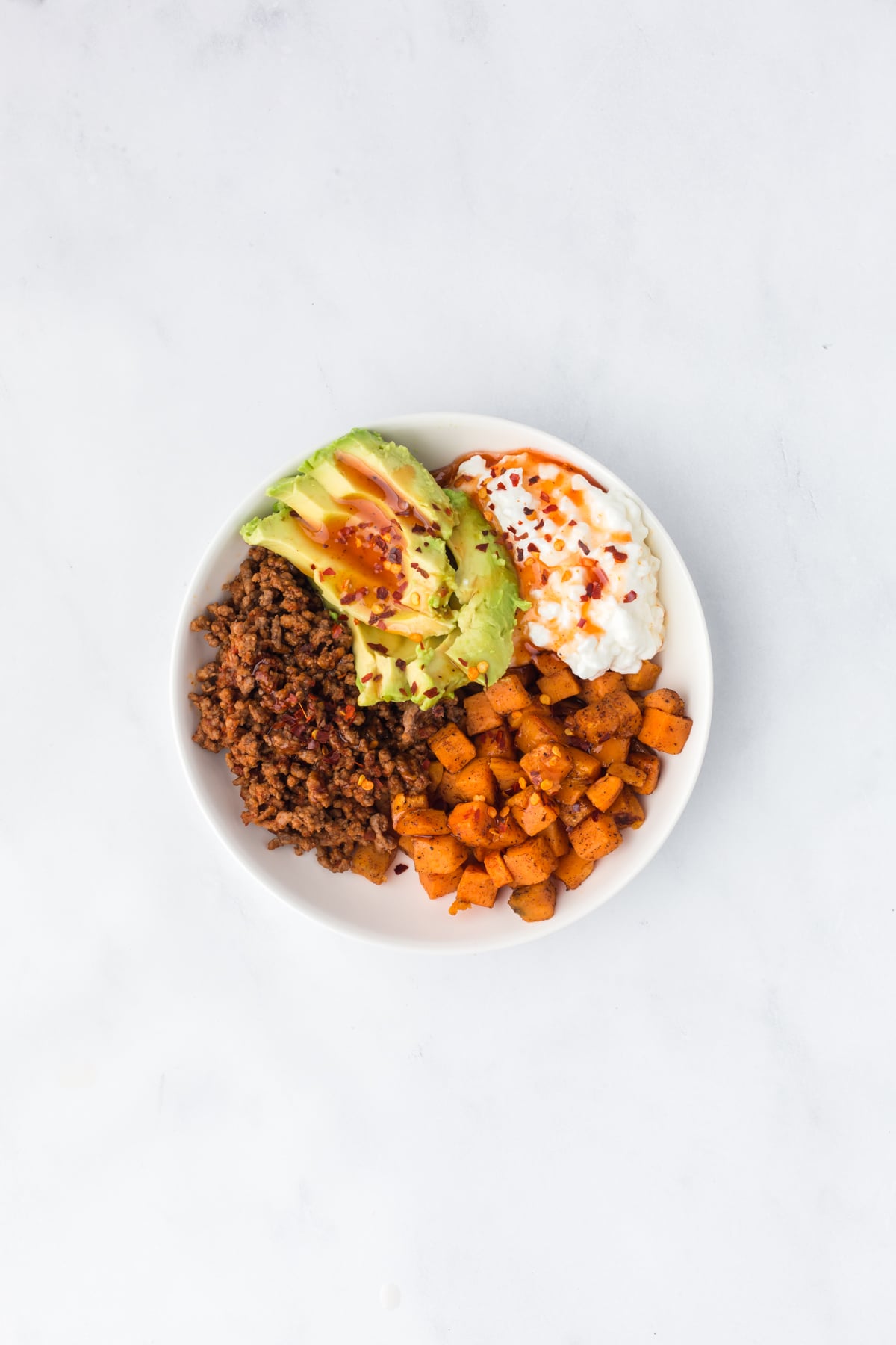 A made bowl filled with cottage cheese, sweet potatoes, ground beef, avocado and a drizzle of hot honey ready to eat.