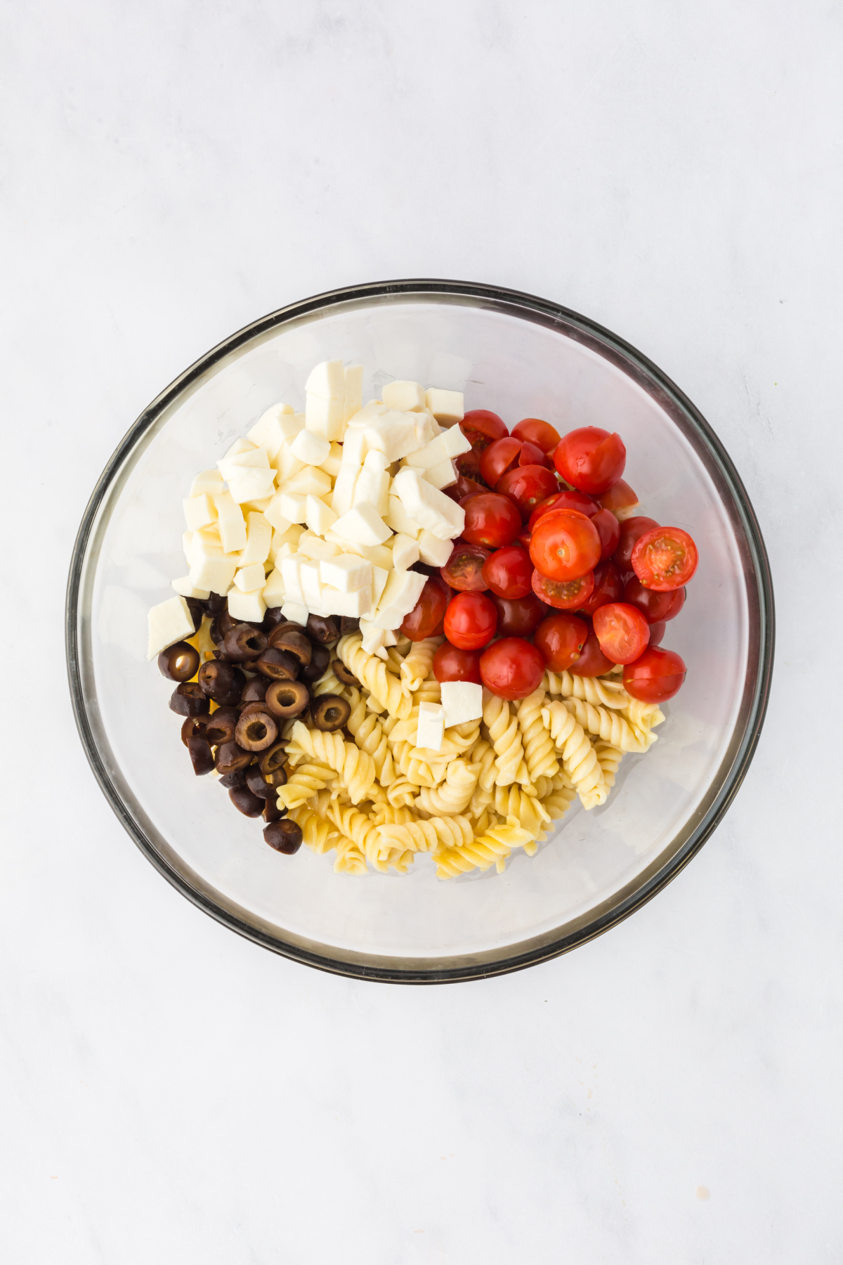 A large bowl full of pasta, cherry tomatoes, mozzarella cubes, and black olives.