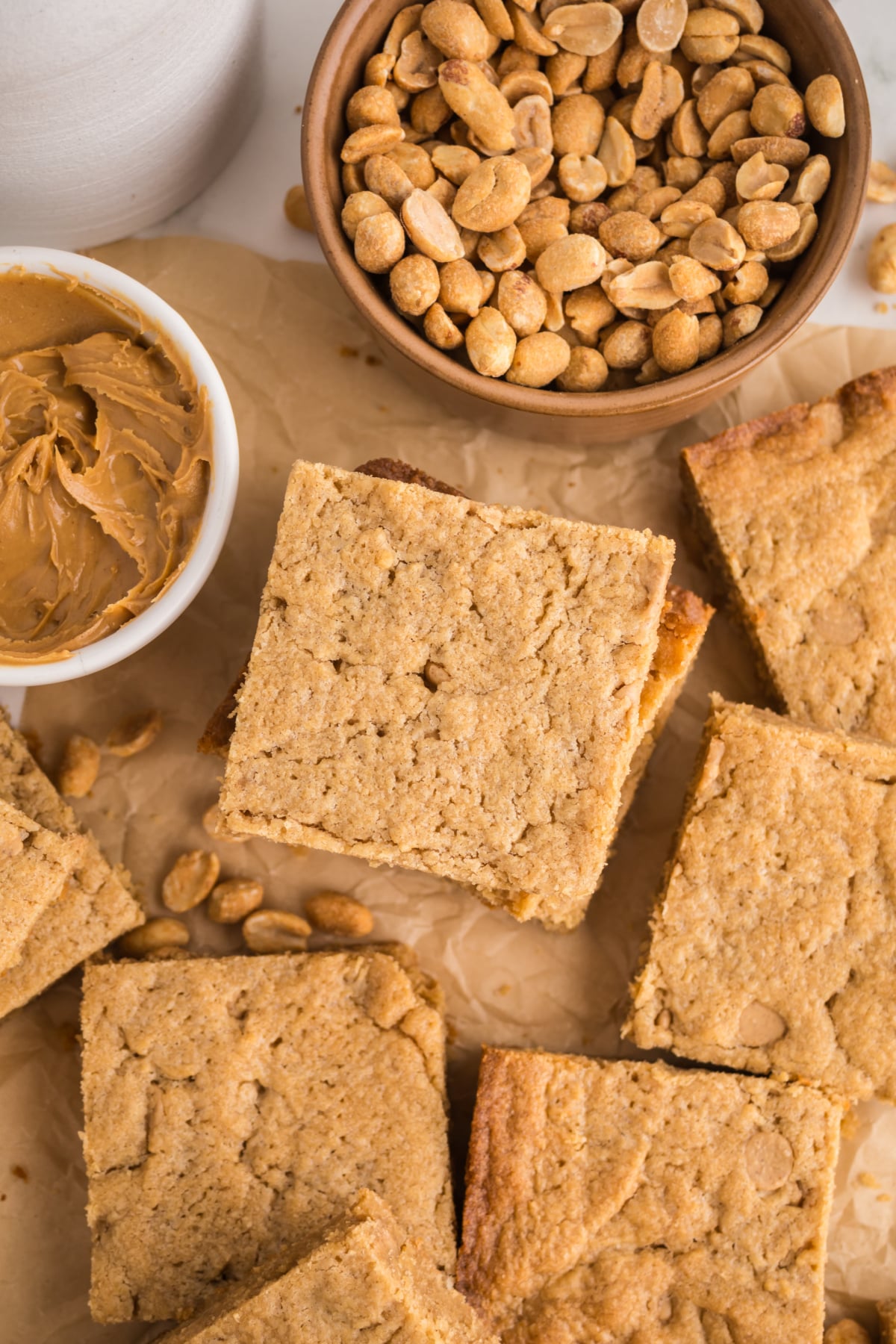 Blondies scattered on parchment with bowl of peanut butter and peanuts.