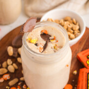 A peanut butter milkshake in a jar with a sprinkling of Reese's pieces and chopped peanut butter cups on top on a table.