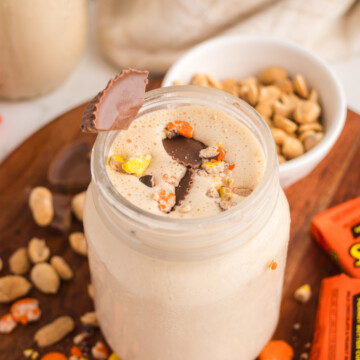 A peanut butter milkshake in a jar with a sprinkling of Reese's pieces and chopped peanut butter cups on top on a table.