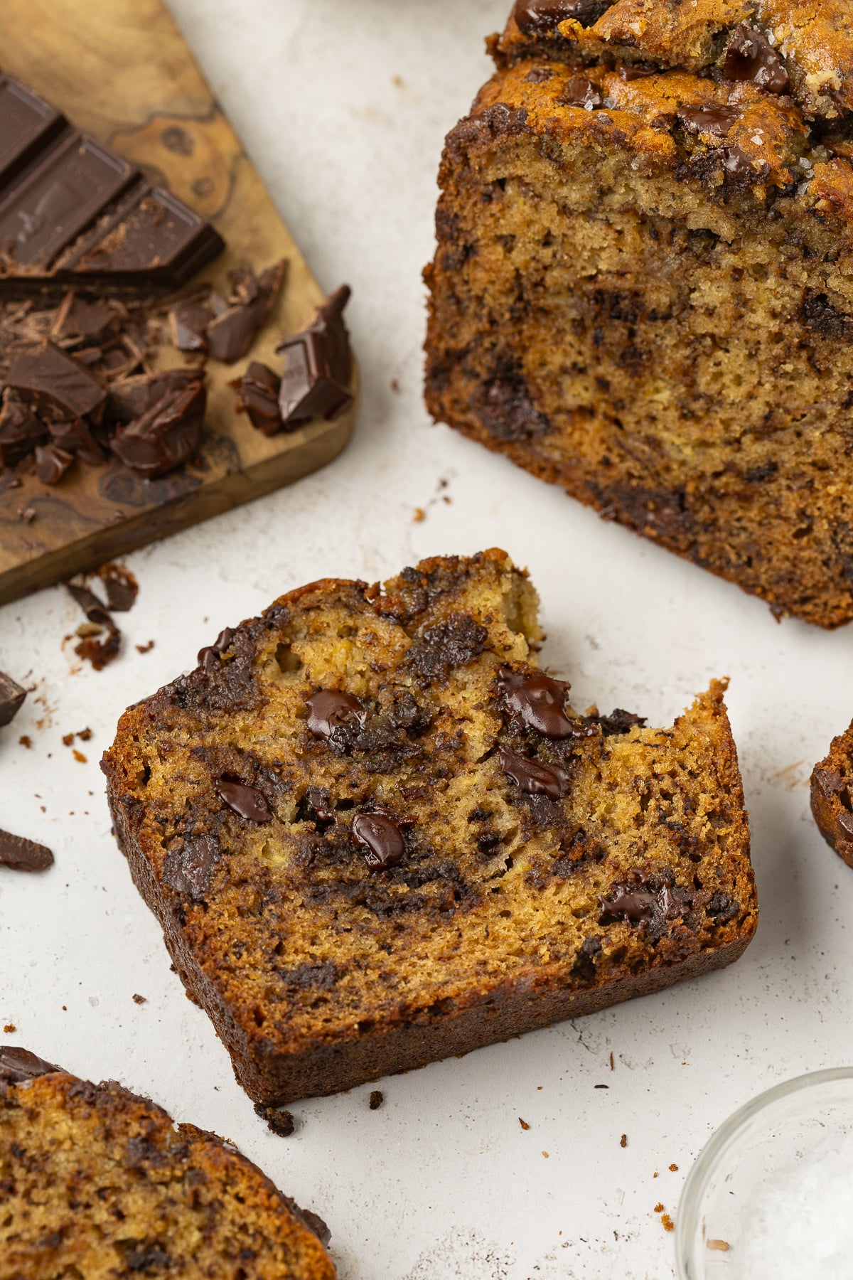 Slice of banana bread missing a bite on counter beside remaining banana bread and some chocolate chunks.