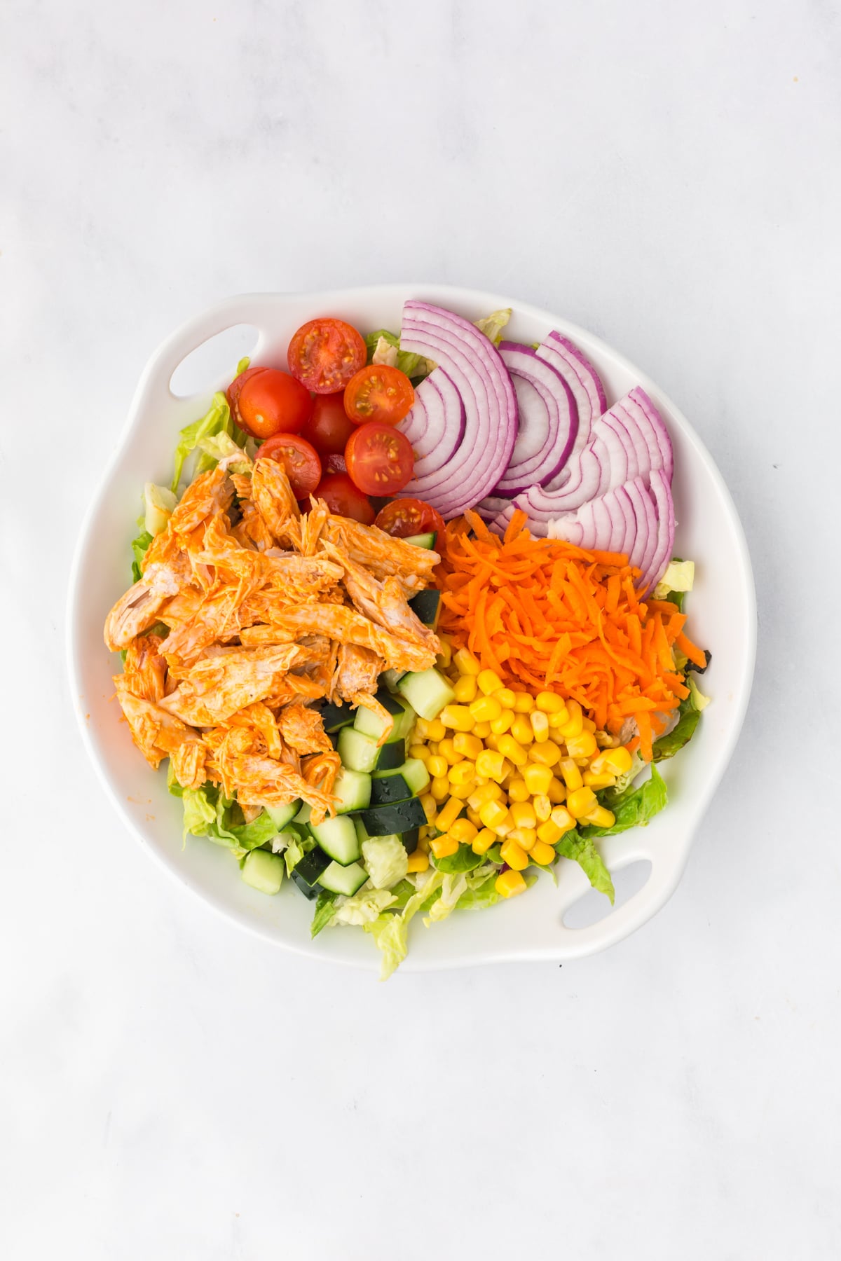 Fresh vegetables and buffalo chicken layered over chopped lettuce in a large salad bowl.
