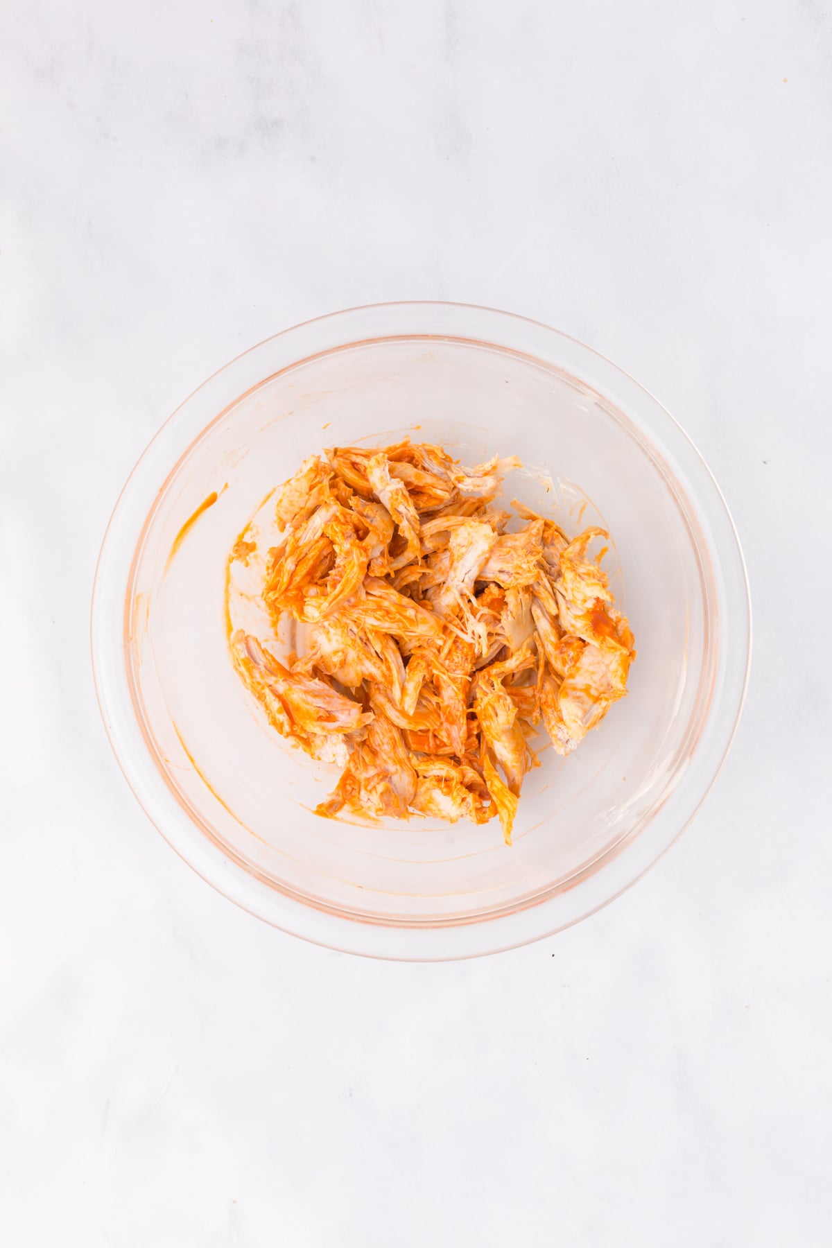 Shredded rotisserie chicken tossed with buffalo sauce in a mixing bowl.